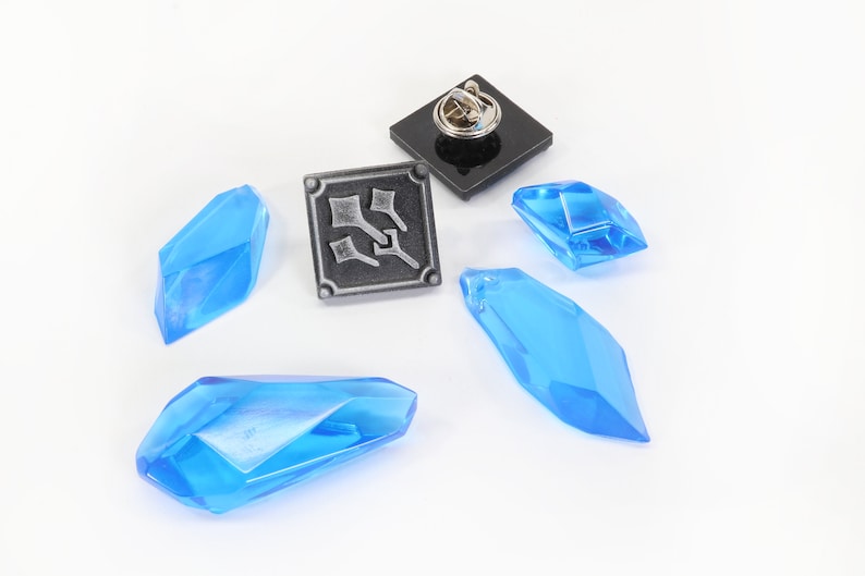 May include: A silver pin with a black background and a design of three swords. There are four blue crystal-like objects on a white background.