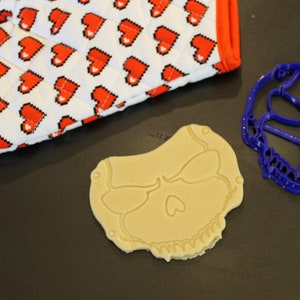May include: A blue plastic skull cookie cutter and a plain, light brown cookie shaped like a skull. The cookie is on a black surface.