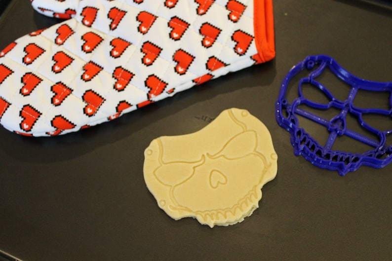 May include: A blue plastic skull cookie cutter and a plain, light brown cookie shaped like a skull. The cookie is on a black surface.