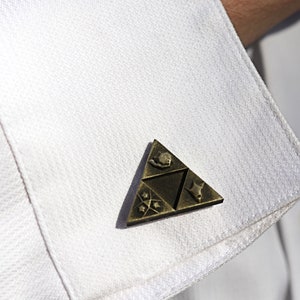 May include: A gold-colored triangular pin with a stylized design. The pin features a triangular shape with four symbols inside. The pin is attached to a white shirt.