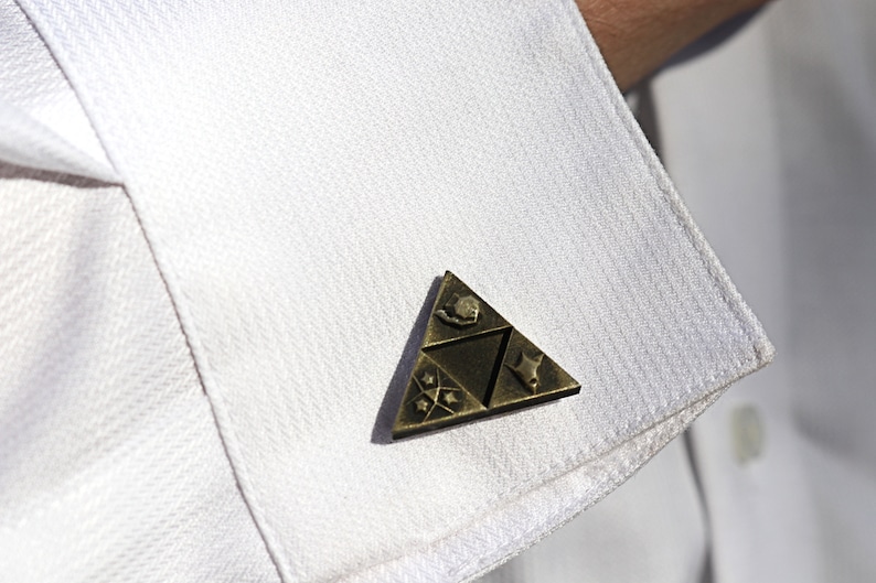 May include: A gold-colored triangular pin with a stylized design. The pin features a triangular shape with four symbols inside. The pin is attached to a white shirt.