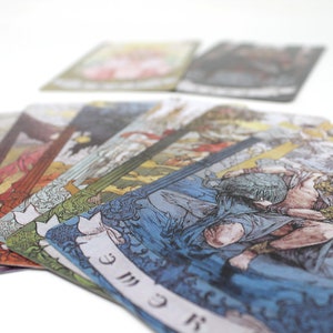 May include: A set of tarot cards with colorful illustrations and gold accents. The cards are fanned out on a white surface, revealing the card "The World" with a woman holding a staff and a wreath.