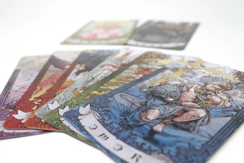 May include: A set of tarot cards with colorful illustrations and gold accents. The cards are fanned out on a white surface, revealing the card "The World" with a woman holding a staff and a wreath.
