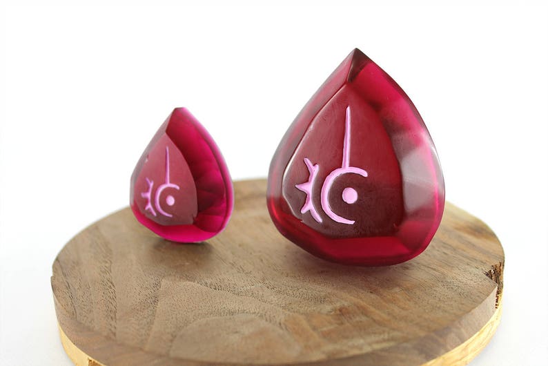 Op de afbeelding: Twee rode glazen hangers in de vorm van een traan met een roze symbool op elk. Het symbool is een halve maan met een zon en een stip in de halve maan.