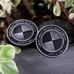 Pu&ograve; includere: Due spille circolari in metallo nero e argento con il testo "Gekkoukan Private High School Since 1982" e un logo stilizzato di un cerchio diviso in quattro quadranti.