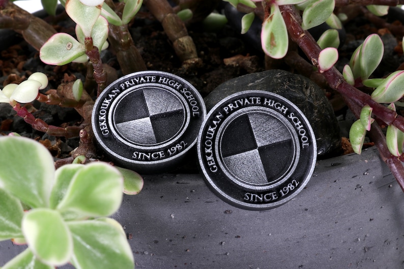 Pu&ograve; includere: Due spille circolari in metallo nero e argento con il testo "Gekkoukan Private High School Since 1982" e un logo stilizzato di un cerchio diviso in quattro quadranti.