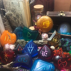 May include: A collection of 12 colorful resin tokens with various symbols and designs. The tokens are arranged on a wooden surface with a book, a candle, and a small wooden chest in the background.