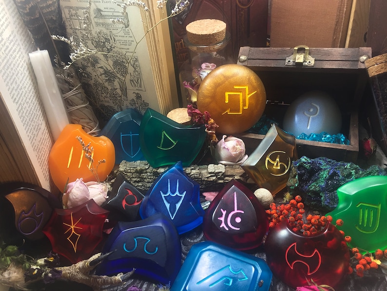 May include: A collection of 12 colorful resin tokens with various symbols and designs. The tokens are arranged on a wooden surface with a book, a candle, and a small wooden chest in the background.