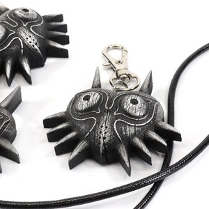 May include: Three silver Majora's Mask pendants, one on a black cord necklace and one on a silver keychain. The masks have a textured, wooden look with sharp, pointed edges.