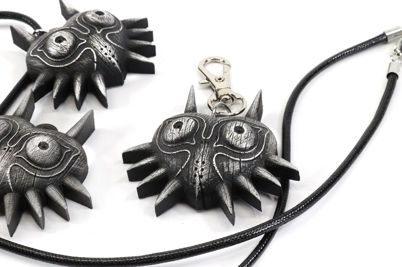 May include: Three silver Majora's Mask pendants, one on a black cord necklace and one on a silver keychain. The masks have a textured, wooden look with sharp, pointed edges.