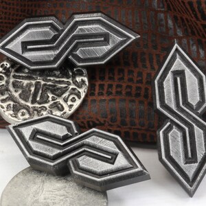 May include: A collection of silver-colored metal pins and a silver button. The pins feature a stylized "S" design with a textured finish and black borders. The button has an embossed design. The background is a brown, textured material.