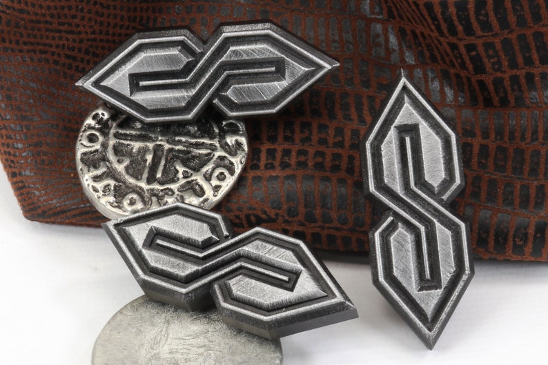 May include: A collection of silver-colored metal pins and a silver button. The pins feature a stylized "S" design with a textured finish and black borders. The button has an embossed design. The background is a brown, textured material.