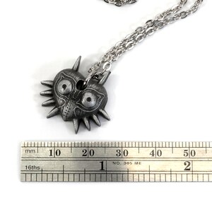 May include: A silver necklace with a Majora's Mask pendant. The pendant is about 2 cm wide. The necklace is on a white background.