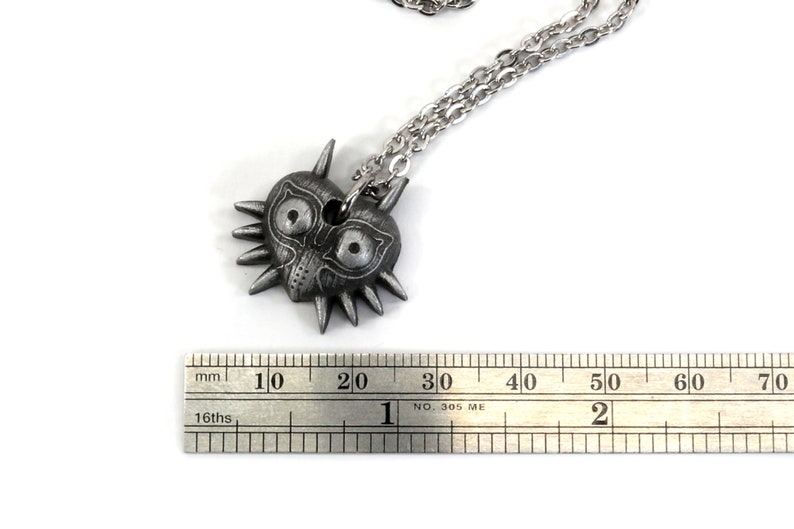 May include: A silver necklace with a Majora's Mask pendant. The pendant is about 2 cm wide. The necklace is on a white background.