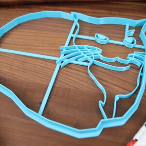 May include: A light blue plastic cookie cutter shaped like a cat's face. The cutter has detailed features, including eyes, a nose, and whiskers.