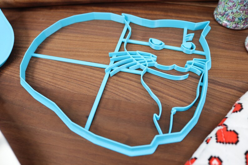 May include: A light blue plastic cookie cutter shaped like a cat's face. The cutter has detailed features, including eyes, a nose, and whiskers.
