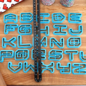 May include: A set of 26 blue plastic alphabet cookie cutters on a wooden surface. The cutters are in a geometric font style.
