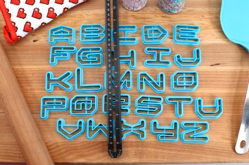 May include: A set of 26 blue plastic alphabet cookie cutters on a wooden surface. The cutters are in a geometric font style.