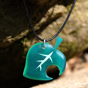 May include: A teal green leaf pendant necklace with a white leaf design and a black cord. The pendant is shaped like a leaf with a hole in the middle.