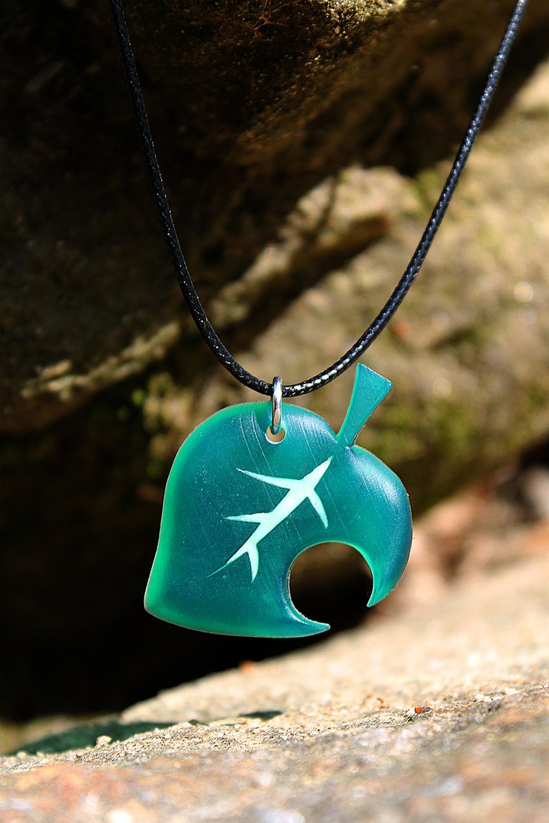 May include: A teal green leaf pendant necklace with a white leaf design and a black cord. The pendant is shaped like a leaf with a hole in the middle.