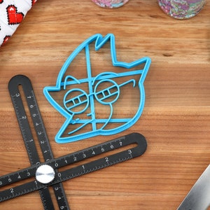 Peut inclure: Un emporte-pi&egrave;ce bleu clair en forme de personnage de dessin anim&eacute; avec des lunettes. Une r&egrave;gle d'angle r&eacute;glable noire, une spatule bleue et un gant de cuisine &agrave; motifs de c&oelig;urs sont &eacute;galement visibles.