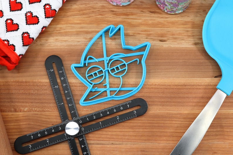 Peut inclure: Un emporte-pi&egrave;ce bleu clair en forme de personnage de dessin anim&eacute; avec des lunettes. Une r&egrave;gle d'angle r&eacute;glable noire, une spatule bleue et un gant de cuisine &agrave; motifs de c&oelig;urs sont &eacute;galement visibles.