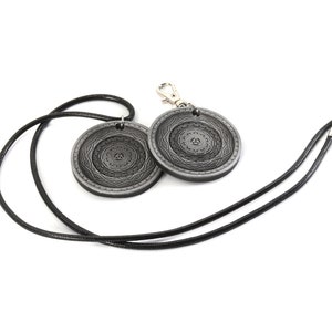 May include: A black leather necklace with two silver pendants featuring intricate circular designs. The pendants have a raised, textured pattern.