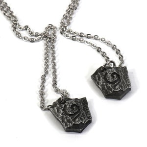 May include: Two silver necklaces with matching pendants. Each pendant is a silver shield with a stylized letter 'G' etched into the surface.