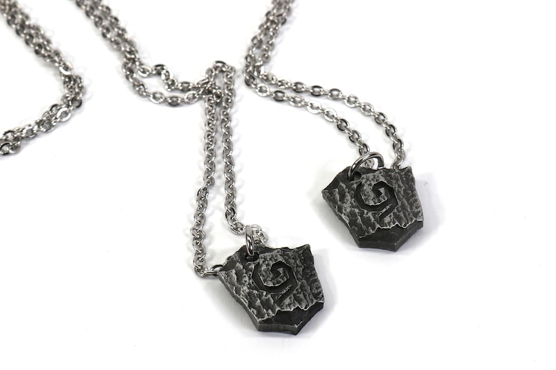 May include: Two silver necklaces with matching pendants. Each pendant is a silver shield with a stylized letter 'G' etched into the surface.