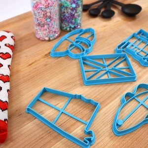May include: A set of blue plastic cookie cutters in various shapes, including a fish, a house, a flag, and a train. The cutters are on a wooden surface with a red and white oven mitt and two jars of sprinkles.