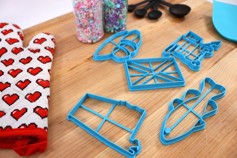May include: A set of blue plastic cookie cutters in various shapes, including a fish, a house, a flag, and a train. The cutters are on a wooden surface with a red and white oven mitt and two jars of sprinkles.
