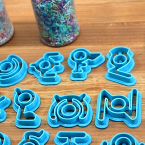May include: A set of 15 blue plastic cookie cutters in various shapes, including circles, swirls, and geometric designs. The cutters are arranged on a wooden surface.