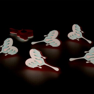 May include: Six heart-shaped, dripping, pastel pink and blue resin ornaments. The ornaments have a glossy finish and are arranged on a black background.
