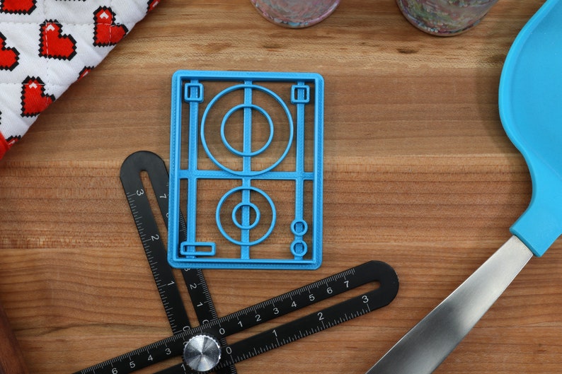 May include: A blue plastic cookie cutter shaped like a soccer field with a circle in the center and two smaller circles on the side.