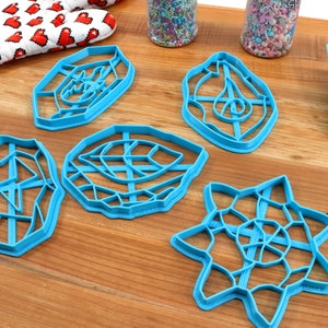 May include: Five blue plastic cookie cutters in different geometric shapes, including a star, a leaf, and a circle. The cutters are on a wooden surface.