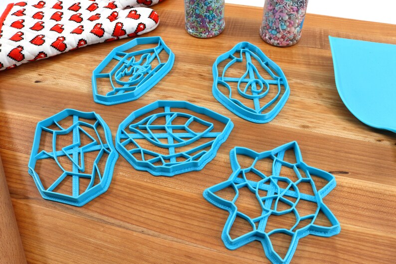 May include: Five blue plastic cookie cutters in different geometric shapes, including a star, a leaf, and a circle. The cutters are on a wooden surface.