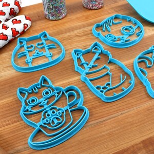 May include: Five blue plastic cat-shaped cookie cutters on a wooden surface. The cutters are in various poses, including sitting, standing, and holding a cup.