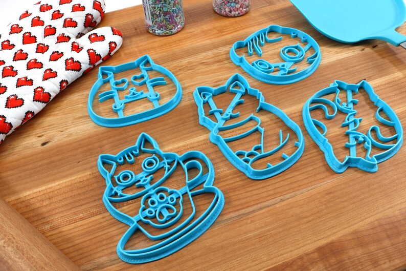 May include: Five blue plastic cat-shaped cookie cutters on a wooden surface. The cutters are in various poses, including sitting, standing, and holding a cup.