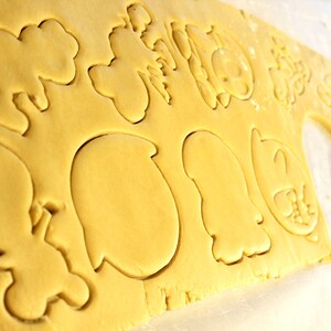 May include: A sheet of yellow cookie dough with various shapes cut out using cookie cutters. The shapes include a cloud, a heart, a star, a circle, a crescent moon, a ghost, and a few other abstract shapes.