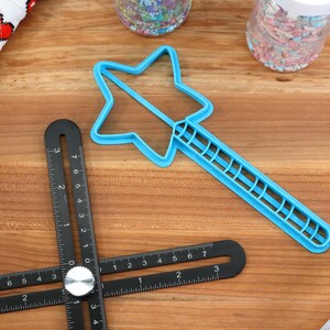 May include: A blue plastic star-shaped cookie cutter with a handle. The cutter is on a wooden surface with a black ruler and a jar of sprinkles.