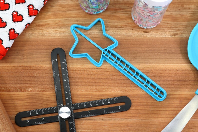 May include: A blue plastic star-shaped cookie cutter with a handle. The cutter is on a wooden surface with a black ruler and a jar of sprinkles.