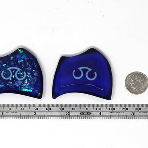 May include: Two blue glass decorative pieces with a white symbol in the center. The piece on the left has a glittery finish. The piece on the right is solid blue.