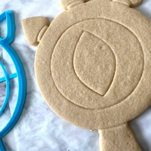 May include: A blue plastic cookie cutter shaped like a target and a plain, unbaked sugar cookie cut into the shape of a shield.
