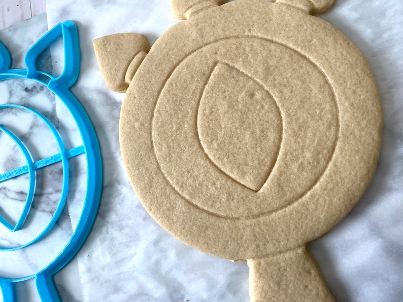 May include: A blue plastic cookie cutter shaped like a target and a plain, unbaked sugar cookie cut into the shape of a shield.