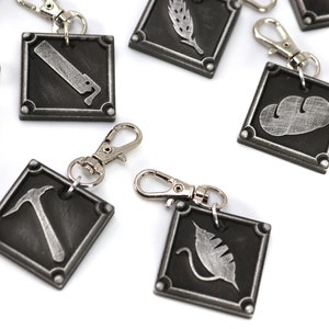 May include: A collection of square, metal charms with various embossed designs, including a saw, wheat, and a cloud. Each charm has a silver-colored clasp for attachment. The charms are dark gray with silver-colored details.