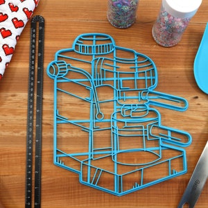 May include: A blue plastic cookie cutter shaped like an espresso machine. The cookie cutter is on a wooden surface with a rolling pin, a ruler, and a spatula.