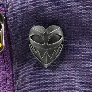 May include: A silver heart-shaped pin with a menacing grin and eyes. The pin is attached to a purple fabric background.