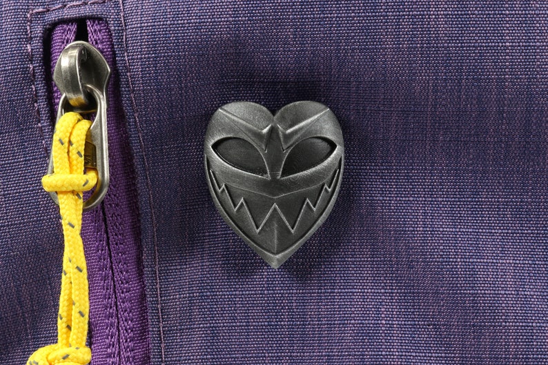 May include: A silver heart-shaped pin with a menacing grin and eyes. The pin is attached to a purple fabric background.