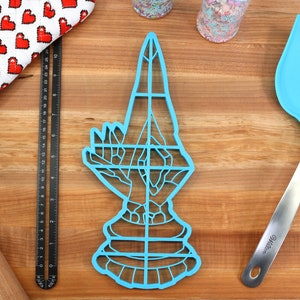 May include: A blue plastic cookie cutter shaped like a hand in a praying position. The cutter has a grid pattern and is on a wooden surface.