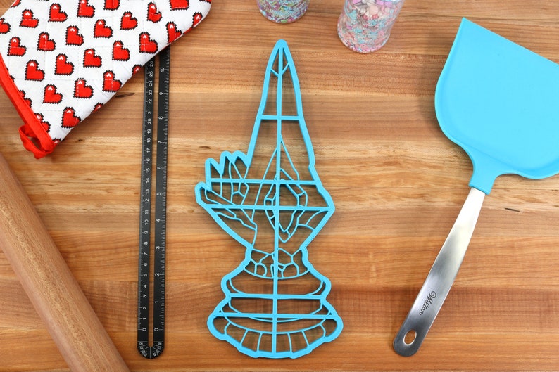 May include: A blue plastic cookie cutter shaped like a hand in a praying position. The cutter has a grid pattern and is on a wooden surface.